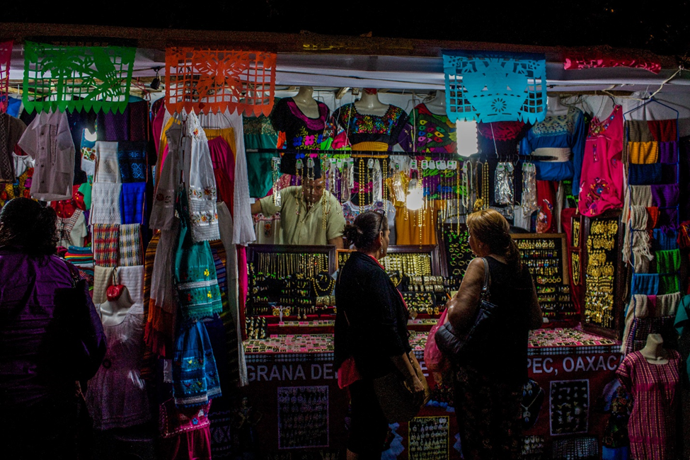 Two women standing in front of a market stall that sells traditional crafts in Oaxaca, Mexico, showcasing a basic form of indigenous involvement in tourism development programs.