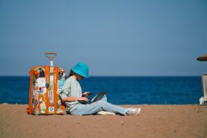 A Gen Z traveler sits on a sandy beach with the ocean in the background. She is leaning against a bright orange suitcase and is working remotely on both her phone and laptop. Photo by Anastasia Nelen on Unsplash.