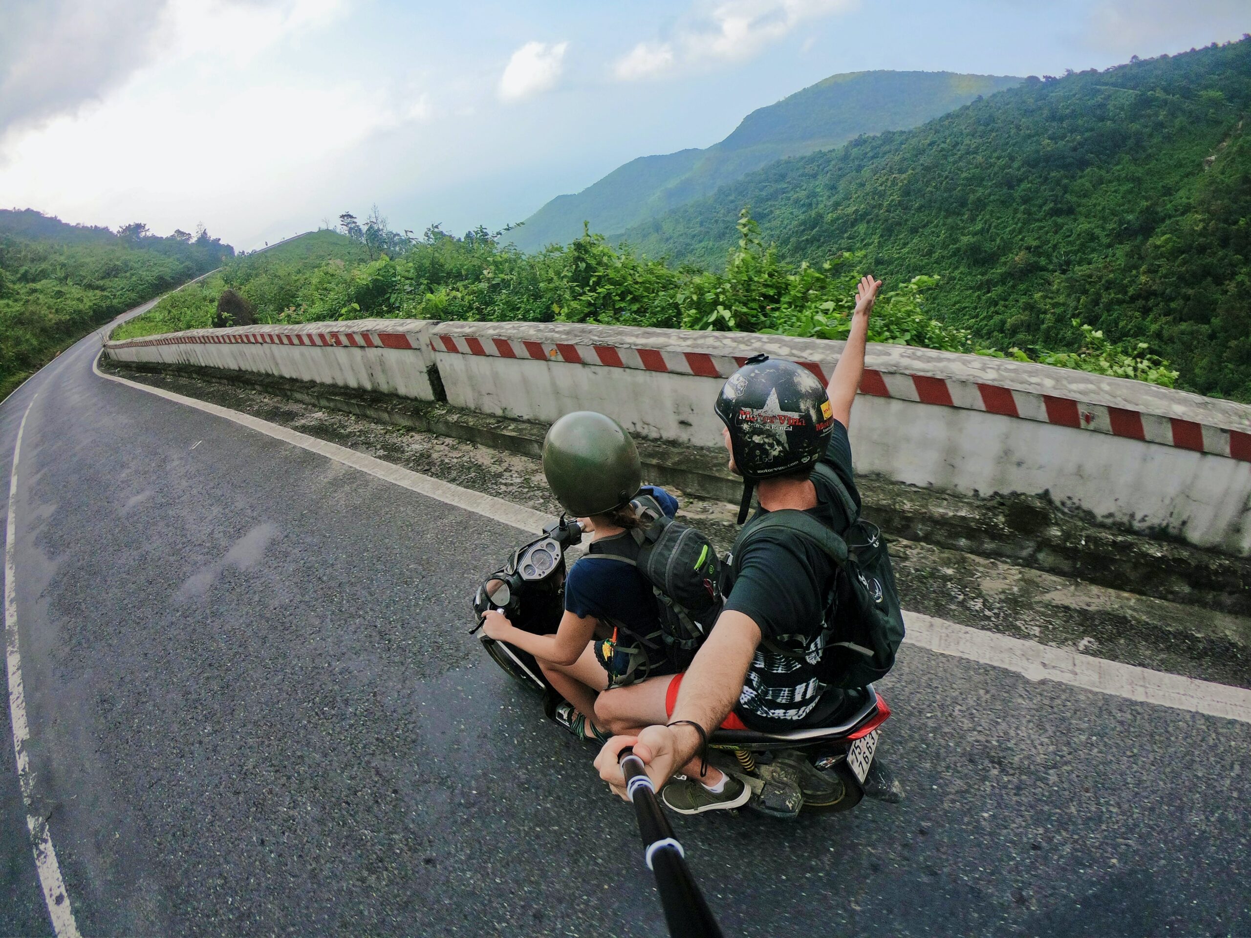 Fish-eye style photo of two Gen Z backpackers riding on a motorbike, a sustainable form of transportation. They are driving on a rural road in central Vietnam, surrounded by rolling hills covered with beautiful green plants. Photo by Jordan Opel on Unsplash.