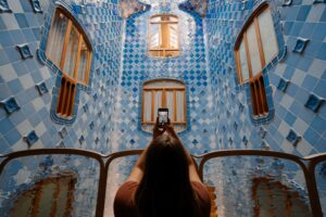 A young Gen Z traveler is taking a photo for her travel blog. She is standing in front of a large wall covered with light and dark blue tiles, with windows letting in lots of natural light, in Barcelona, Spain. Photo by Karsten Winegeart on Unsplash.