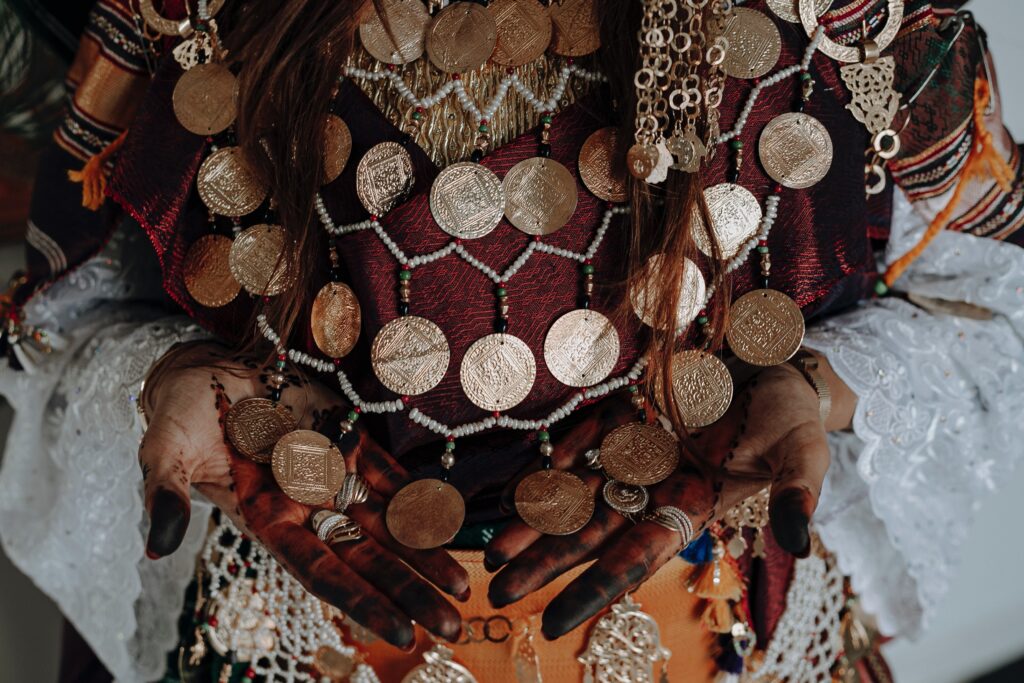 Traditional Tunisian woman wearing stunning gold pendant and bead necklace, vibrant top with traditional patterns, intricate henna designs on hands, matching beaded belt with gold pendants, and rings.